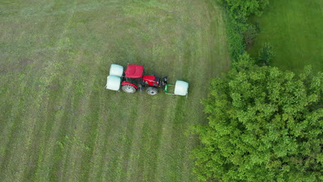 Farmer-in-red-tractor-picking-up-grass-bales-on-meadow,-wide-angle-aerial-view,-nobody