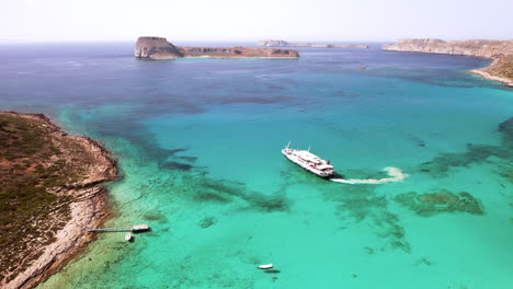 Aerial-Tracking-Shot-of-Day-Cruise-Boat-Departing-Balos-Beach-in-Crete,-Greece-in-Beautiful-Turquoise-Water-on-Sunny-Day