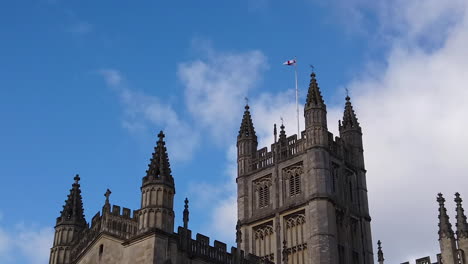 Shot-of-Seagull-Flying-in-and-out-of-Frame-with-Tower-of-Bath-Abbey-in-Somerset,-England-in-the-Background-on-Sunny-Summer’s-Day-with-Saint-George’s-Cross-Flapping-in-the-Wind