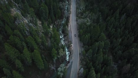 Cinematic-and-beautiful-drone-shot-panning-up-and-following-a-car-driving-through-a-canyon-in-the-Rocky-Mountains,-surrounded-by-pine-trees-on-both-sides-of-the-road