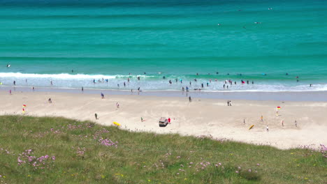 Ocean-waves-along-the-wide-sandy-shore-at-Sennen-Cove-in-Cornwall-with-surfers-and-tourists-walking-on-the-beach