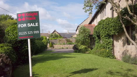Static-Shot-of-British-‘For-Sale’-Real-Estate-Sign-on-Sunny-Summer’s-Day-with-Rural-Village-Home-in-Background