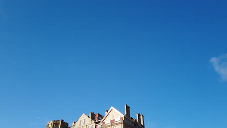 Pulteney-Weir-and-the-Empire-Hotel-in-Bath,-Somerset-on-a-Beautiful-Summer’s-Morning-fading-in-diagonally-from-Clear-Blue-Sky-with-Seagull-Flying-Across-Frame-in-Slow-Motion