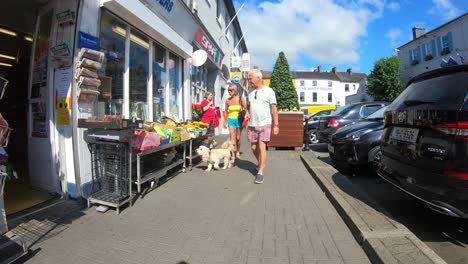 Handheld-smooth-footage-of-a-summer-street-in-a-small-town-with-dog-walkers-passing-by-a-small-shop