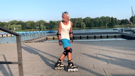 Cheerful-old-fit-senior-man-is-riding-backwards-on-rollerblades-at-lake-during-sunset