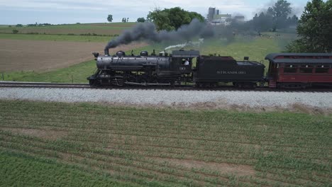 Vista-Aérea-De-Un-Motor-De-Vapor-Antiguo-Con-Automóviles-De-Pasajeros-Resoplando-A-Lo-Largo-De-La-Campiña-Amish