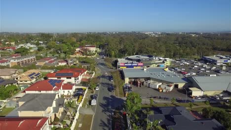 Aerial-drone-view-along-a-suburban-road,-past-a-local-shopping-centre-towards-a-busy-arterial-road