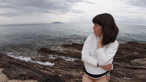 A-brunette-girl-sitting-alone-on-a-rocky-beach-on-the-Mediterranean-on-a-partly-cloudy-day,-with-an-island-in-the-background