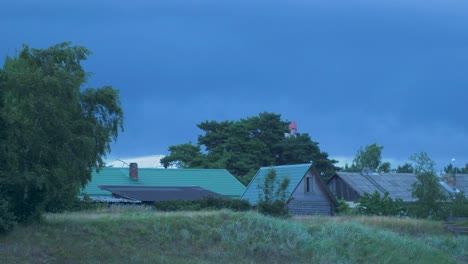 Fast-moving-dark-gray-rain-clouds-over-the-wooden-buildings-and-port-navigation-beacon-with-red-light-in-evening,-wide-shot
