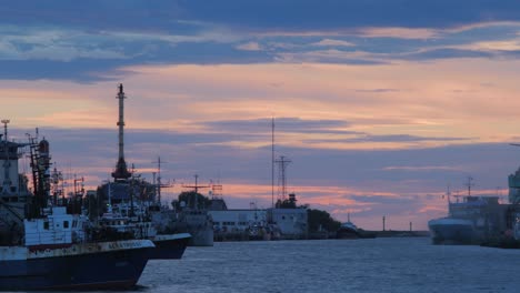 Lapso-De-Tiempo-Con-Hermosas-Nubes-Escénicas-De-Puesta-De-Sol-Moviéndose-Sobre-El-Canal-Comercial-Con-Barcos-Y-Grúas-Portuarias-En-Funcionamiento-En-El-Puerto-De-Liepaja,-Plano-General