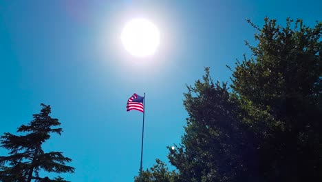 Shot-of-US-Flag-waving-in-the-wind-on-a-flag-pole-amidst-trees-with-the-sun-shining-behind-it-in-the-city-of-Solvang,-California,-USA