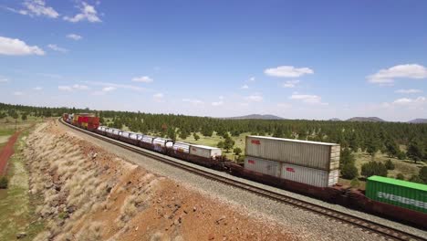 An-aerial-of-cargo-train-pulling-container​-cars,-oil-cars,-and-flatbed,-passes-close-to-the-camera-as-it-heads-west