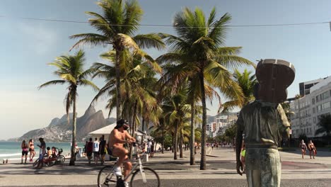 Ipanema-Mit-Bronzestatue-Des-Komponisten-Tom-Jobim,-Der-Eine-Gitarre-über-Der-Schulter-Hält,-Mit-Palmen-Im-Hintergrund-Und-Vorbeigehenden-Menschen,-Die-Zu-Fuß-Gehen-Und-Radfahren