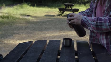 Woman-pouring-drink-from-flask-at-wooden-picnic-table-medium-shot