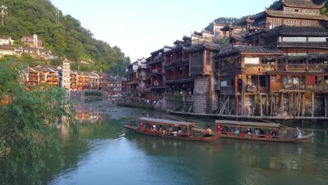 Hacer-Turismo-En-Viejos-Barcos-Turísticos-De-Madera-Navegando-Entre-Las-Antiguas-Casas-De-Madera-Sobre-Pilotes-Construidas-A-Orillas-Del-Río-Tuo,-Que-Fluyen-Por-El-Centro-Del-Casco-Antiguo-De-Fenghuang