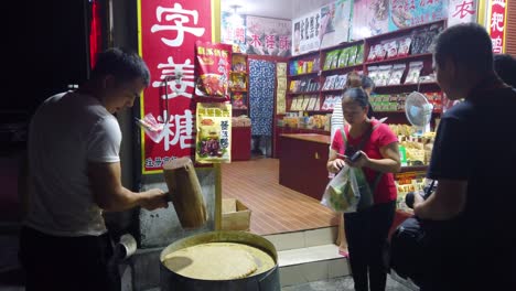 Tourists-looking-at-a-chinese-man-hitting-hard-with-big-wooden-hammers-to-crack-grain-which-will-be-used-in-making-of-sweet-snack-for-sale-on-the-street,-China