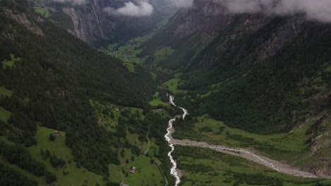 An-drone-shot-moving-forwards-and-tilting-up,-revealing-a-small-creek-running-down-from-the-mountain-to-the-small-village,-surrounded-by-beautiful-and-lush-greenery-in-the-summer-in-Lauterbrunnen