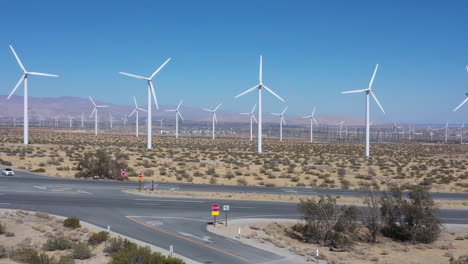 Renewable-onshore-turbines-in-wind-farm-generating-green-energy-by-countryside-expressway-and-traffic-with-on-blue-cloudless-sunny-sky-day,-aerial-sideways