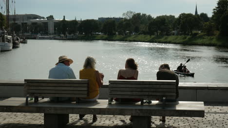 Rear-shot-of-family-sitting-on-wooden-bench,enjoying-beautiful-river-view-during-sunny-day