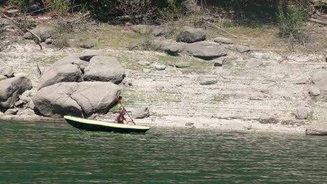 A-Kayaker-Paddling-On-The-Calm-River-On-A-Sunny-Day,-Rocky-Riverbank-In-The-Background---wide-shot