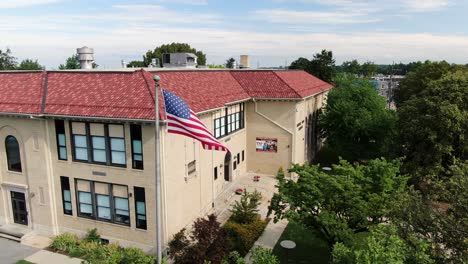 Beautiful-aerial-push-in-on-American-flag-in-breeze,-two-story-public-school-building,-Spanish-architecture,-establishing-shot,-education-theme