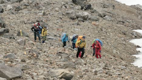 Determined-adult-men-and-women-wearing-bright-full-hiking-gear,-walking-through-rocky-path,-looking-up-vast,-snowy-mountain,-Mt