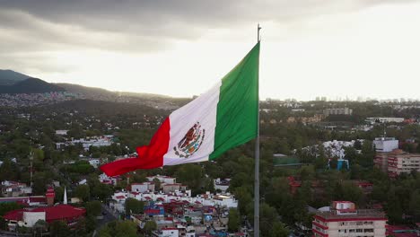 Flight-by-large-majestic-patriotic-red,-white-and-green-Mexican-flag-waving-in-wind-in-downtown-Mexico-city-center,-San-Jeronimo-neighborhood-on-overcast-cloudy-grey-sky-day,-sideways-aerial