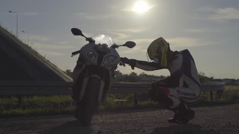 Medium-closeup-shot-of-a-biker-in-full-body-suit-wearing-helmet-squatting-and-praying-next-to-a-motorcycle-with-the-sun-and-a-bridge-in-the-background