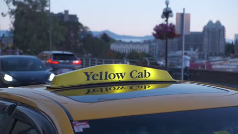 Freihandaufnahme-Des-Dachs-Eines-Gelben-Taxis-Mit-Beleuchtetem-Schild-Und-Vorbeifahrendem-Nahverkehr-Im-Hintergrund-An-Einem-Warmen-Sommerabend-In-Victoria,-BC