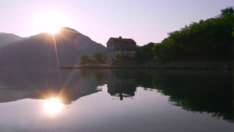 Die-Sonne,-Die-Hinter-Einer-Bergkette-über-Der-Bucht-Von-Kotor,-Montenegro,-Aufgeht,-Ihr-Licht-Reflektiert-Sich-Im-Wasser-Und-Scheint-Auf-Ein-Herrenhaus-Und-Eine-Kleine-Küstenstadt-An-Seinen-Ufern,-Luftaufnahme