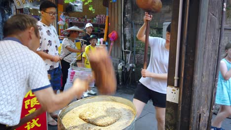 Man-and-woman-hitting-hard-with-big-wooden-hammers-to-crack-grain-which-will-be-used-in-making-of-sweet-snack-for-sale-on-the-street,-China