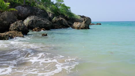 Schöne-Niedrige-Drohne,-Die-über-Das-Klare-Blaue-Atlantikwasser-Des-Tropischen-Strandes-Playa-Preciosa-Fliegt,-Mit-Meeresfelsen,-Palmen-Und-Grünen-Sträuchern-Im-Hintergrund,-Dominikanische-Republik