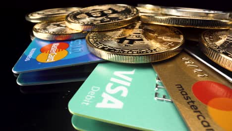 Makroansicht-Goldene-Bitcoins-Münzen-Und-Bank-Debit-Kreditkarten-Von-Mastercard-Visa,-Die-Sich-Auf-Reflektierender-Schwarzer-Glasoberfläche-Umdrehen,-Kryptowährungsinvestition-4k