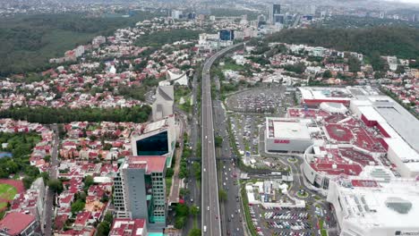 High-aerial-flight-above-car-traffic-traveling-on-Mexico-City-Periferico-Sur-expressway-by-capital-city-center-shopping-mall,-skyscrapers,-and-buildings-on-hazy-day,-Mexico,-overhead-drone-approach
