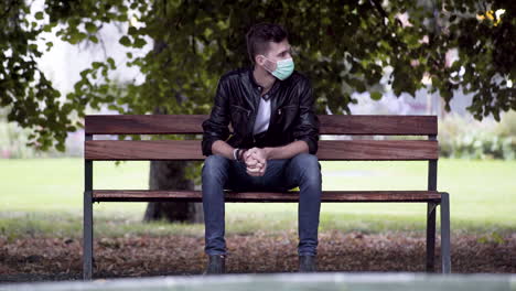 A-young-cool-man-in-jeans,-white-T-shirt-and-a-black-leatherette-jacket,-wearing-a-green-Covid-19-facemask,-sitting-on-a-park-bench-under-a-tree-beside-a-fountain,-impatiently-waiting-for-someone,-4k