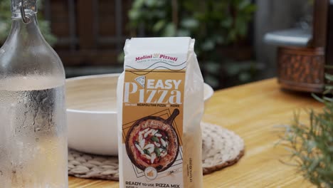 Close-up-on-pizza-flour-package-on-a-wooden-cooking-table-with-pizza-making-oven-in-background