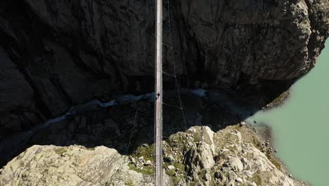 A-static,-symmetrical-shot-of-a-solo,-young-man-is-walking-alone-by-himself-across-Trifthutte-in-Switzerland-on-a-suspension-bridge,-in-perfect-center-and-middle-above-a-small-river-or-stream