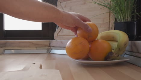 Die-Hand-Des-Mannes-Greift-Nach-Orangenfrüchten-Auf-Einem-Teller-Mit-Banane-Und-Wird-Auf-Ein-Schneidebrett-Gelegt