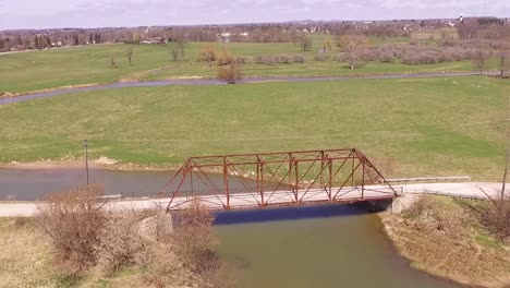 Aerial-over-rural-road,-river-and-steel-bridge