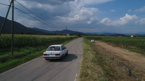 Bmw-E30-Clásico-Blanco-Conduciendo-Cerca-De-Campos-De-Maíz-En-Una-Carretera-Rural-Cerca-De-Hostalets-De-Bas,-Cataluña,-España