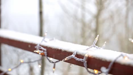 Close-up-of-white-Christmas-lights-on-a-snowy-deck-with-aspen-trees-in-the-background-and-snow-falling