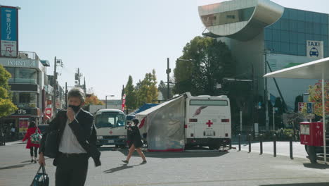 People-Wearing-Face-Mask-In-Street-Of-Tokyo-During-The-Pandemic-COVID-19-With-Red-Cross-Station-In-Background