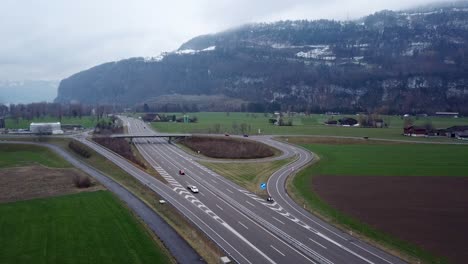 Vista-Aérea-De-Los-Coches-Que-Circulan-Por-La-Autopista-Suiza-Durante-El-Día-Nublado-Con-Montañas-En-El-Fondo-Cubiertas-De-Nieve