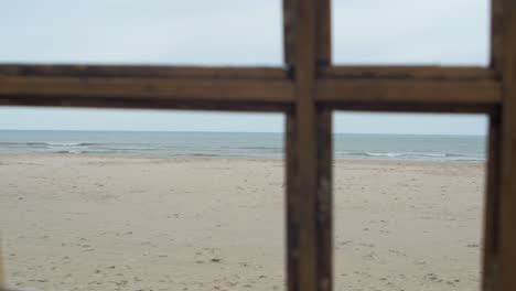 View-of-the-Baltic-sea-with-white-dunes-trough-the-old-wooden-window,-concept,-overcast-cloudy-day,-handheld-shot