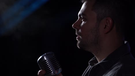 Male-singer-with-unidyne-elvis-vintage-microphone-in-hand-breathing-visible-air-and-turning-head-slowling-up-about-to-sing-at-dark-black-abandoned-industrial-place