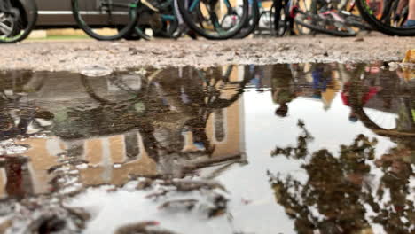 Reflection-of-people-on-bicycles-in-puddle-with-raindrops,-slow-panning-shot-right-to-left