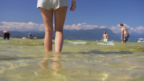 Medium-shot-of-a-girl,-a-woman-walking-through-shallow-lake-water,-on-Jamaica-Beach,-Sirmione,-Lago-Garda-Lake-Garda,-Italy
