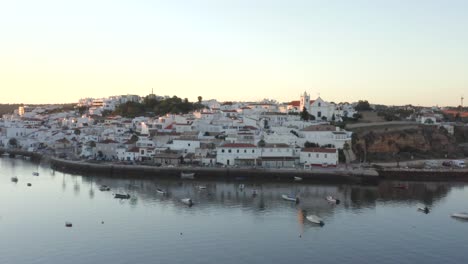 A-calm-river-flowing-in-front-of-beautiful-white-houses-as-the-sun-casts-light-on-rooftops-in-Ferragudo,-Portugal