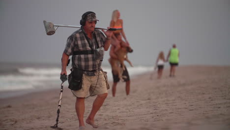 Man-using-metal-detector-on-beach-with-young-couple-in-background,-Slow-Motion