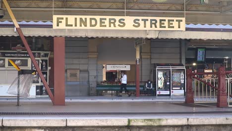 Ein-Zug-Fährt-Während-Des-Ausbruchs-Und-Der-Sperrung-Des-Australischen-Coronavirus-Am-Bahnhof-Flinders-Street-In-Melbourne-An-Maskierten-Passagieren-Vorbei